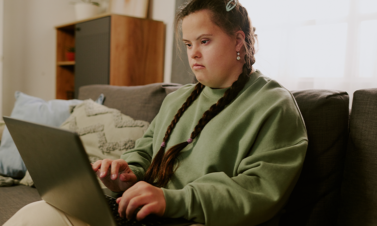 A woman with braided hair sits on a couch using a laptop, looking at the screen as she types.