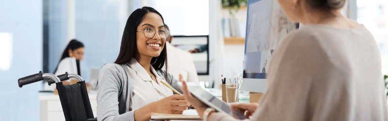 A person using a wheelchair smiles while speaking with another person across a desk in a bright office setting.
