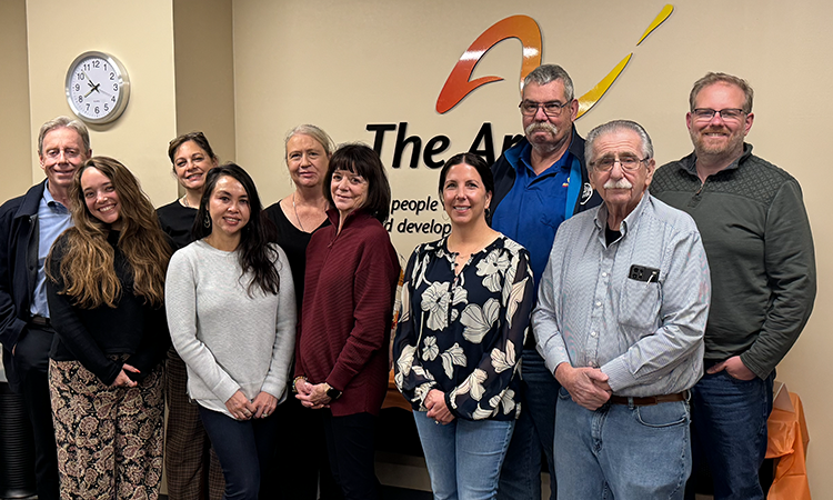A group of ten people stand together indoors, posing in front of a wall with âThe Arcâ logo. A clock and office wall are visible behind them.