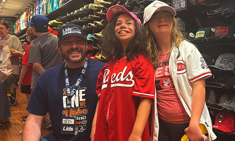 Three people wearing Cincinnati Reds apparel smile inside a sports merchandise store; the manâs cap reads âLids Foundation.â