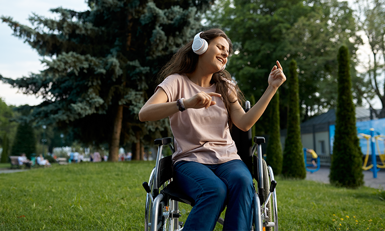 A person in a wheelchair wearing headphones smiles and dances with hands raised while sitting outside in a grassy park area