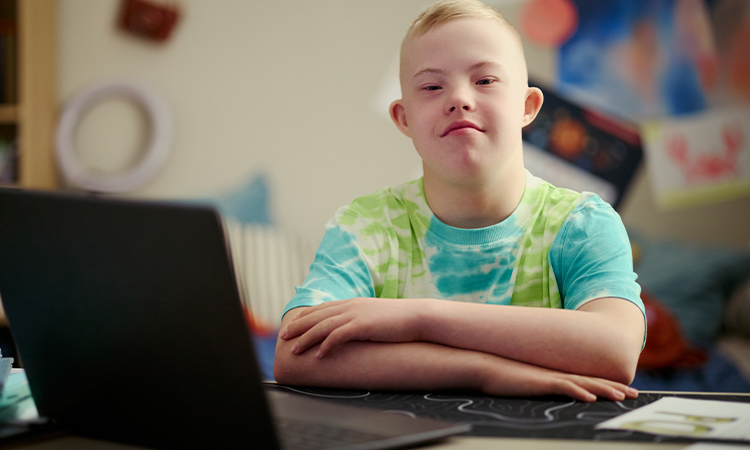 Young person seated at a desk with arms folded, facing the camera beside an open laptop.