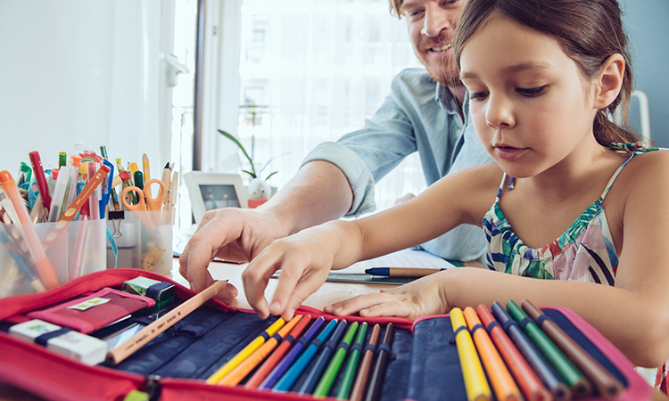 A child selects colored pencils from an open art-supply case while an adult assists at a table.