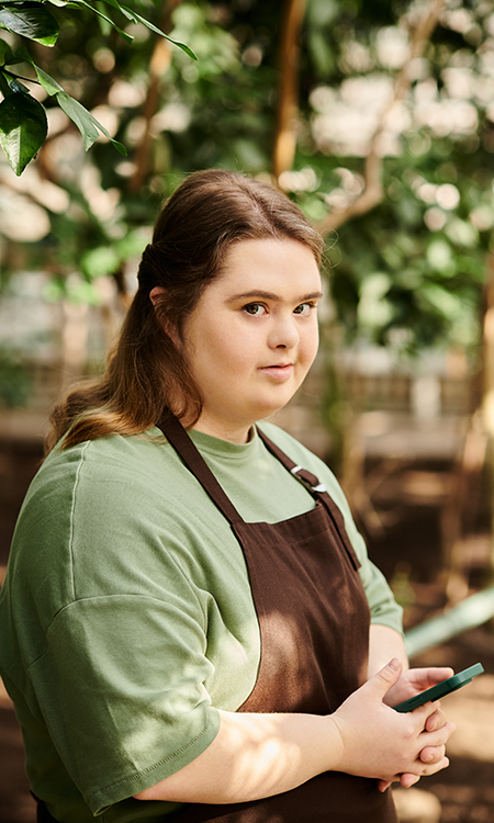 A young woman in a green shirt and brown apron holds a phone while standing among plants in a garden setting.