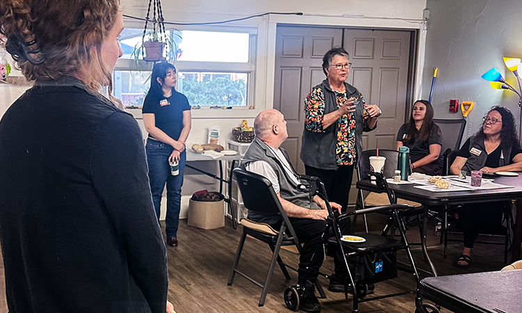 A person stands and speaks to a small group seated around a table while another person sits using a walker nearby; others watch and listen.