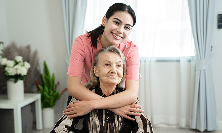 A caregiver stands behind an older woman seated in a chair, smiling with arms gently around her shoulders in a home setting.