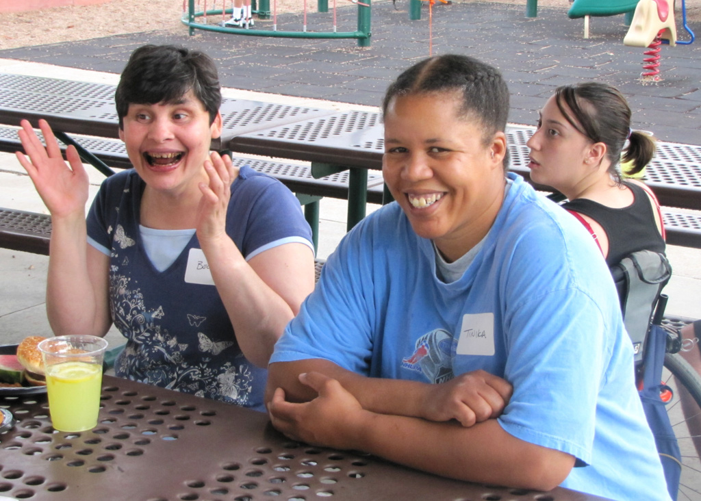 Three people sit together at a picnic table near a playground, with two smiling toward the camera and one person using a wheelchair in the background.