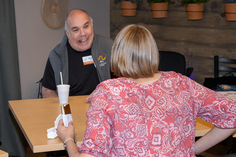 A man wearing a vest with The Arc logo smiles while talking to a woman in a pink patterned shirt at a table indoors