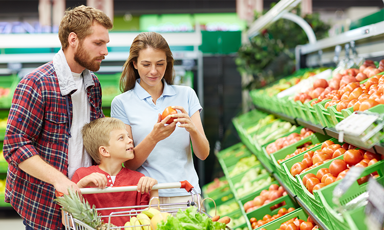 A family shops in a grocery store produce section, with one adult holding a tomato and a child sitting in the cart. Bins of tomatoes and other fresh produce line the aisle.