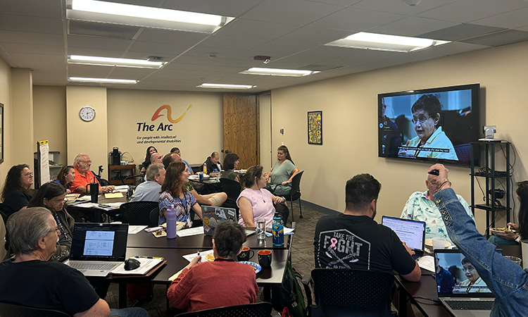 A group of people seated around tables in a meeting room, using laptops and watching a person speaking on a wall-mounted screen. The Arc logo is visible on the wall.