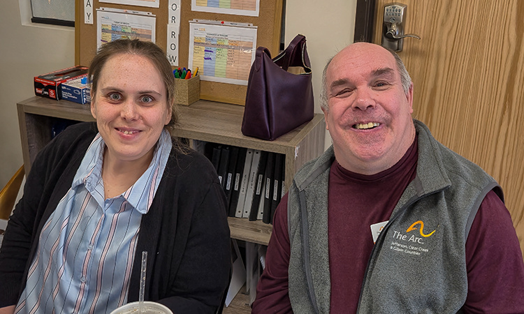 Two people sit side by side at a desk, smiling at the camera in an office setting. Behind them are binders and a bulletin board with papers; one person wears a vest with âThe Arc Jefferson, Clear Creek & Gilpin Countiesâ logo.