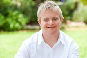 A smiling young man with short blond hair and a white collared shirt sits outdoors in a grassy setting.