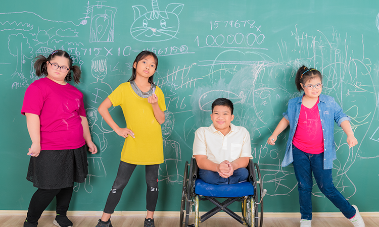 Four children pose in front of a classroom chalkboard covered with drawings and numbers; one child uses a wheelchair.