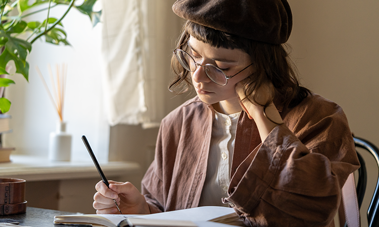 A person wearing glasses and a beret sits by a window writing in a notebook at a desk with soft natural light.