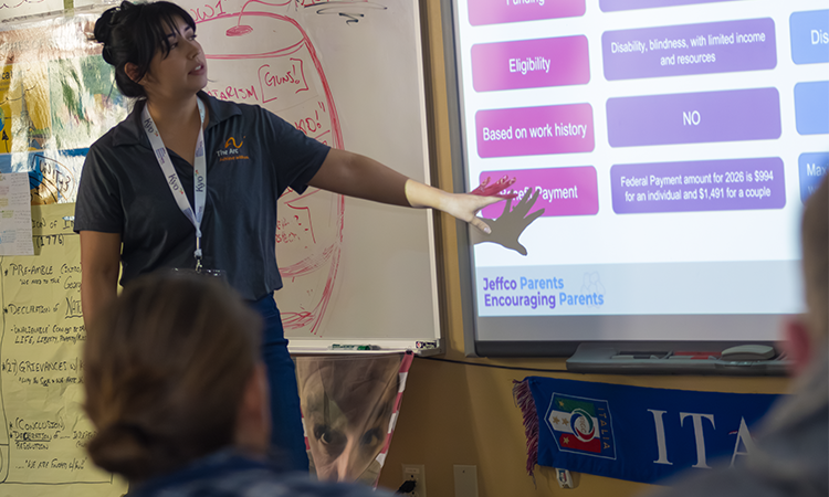 A person points to a projected presentation slide in a classroom while speaking to an audience. Text on the slide includes âEligibility,â âBased on work history,â âNO,â and âMonthly Payment,â along with smaller explanatory text.