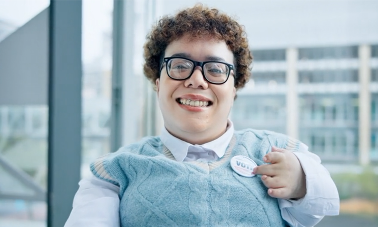A person wearing glasses smiles and points to a button on their shirt that reads â€œVOTE.â€