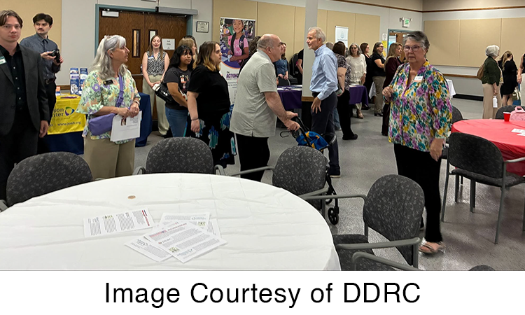 People gather and talk at a community event in a meeting room with tables, chairs, and informational displays. Text reads, â€œImage Courtesy of DDRC.â€