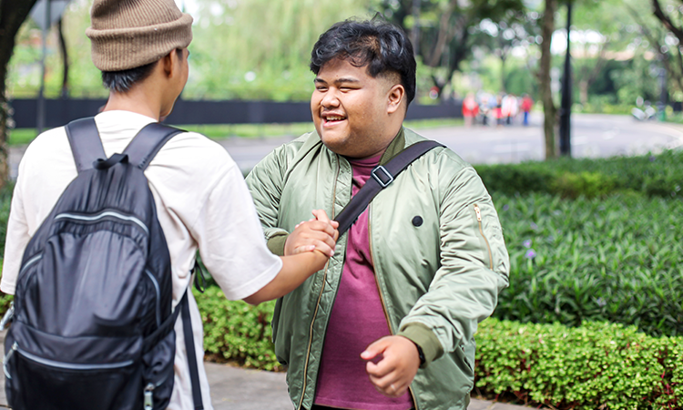 Two young adults greet each other with a handshake outdoors on a tree-lined walkway; one wears a backpack and knit cap, while the other smiles.