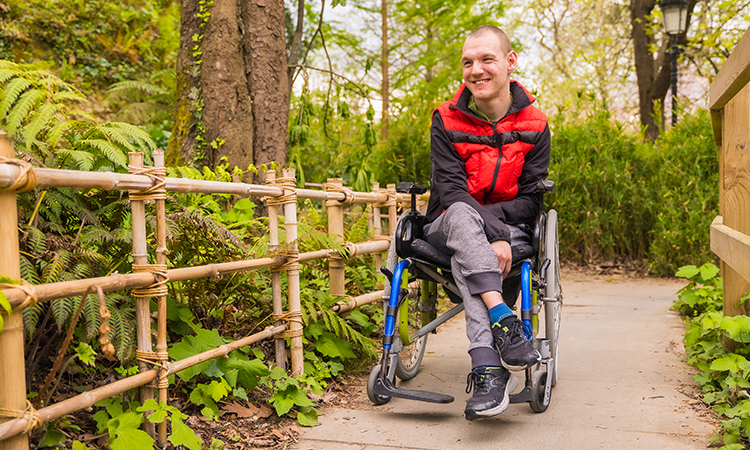 A person using a wheelchair moves along a paved path in a green park, smiling. A wooden fence and trees line the path.
