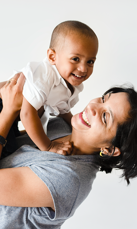 An adult smiles while lifting a young child in the air, both appearing joyful against a plain background.
