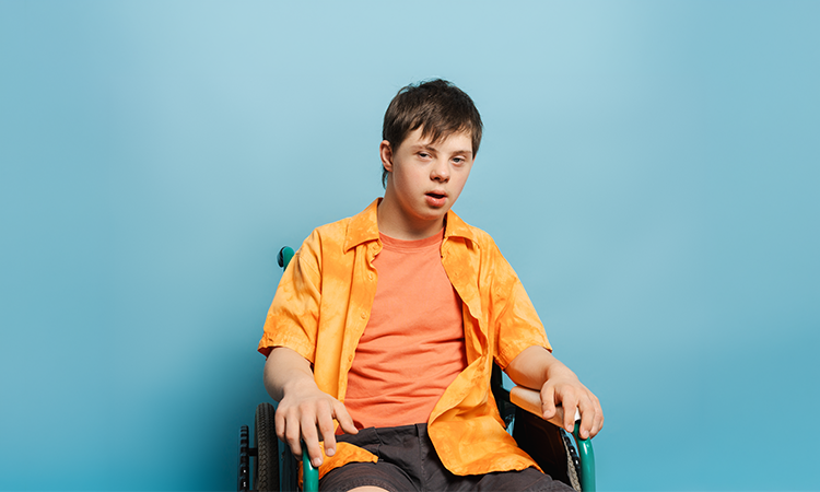 A teenager using a wheelchair wears an orange shirt over a coral T-shirt against a light blue background.