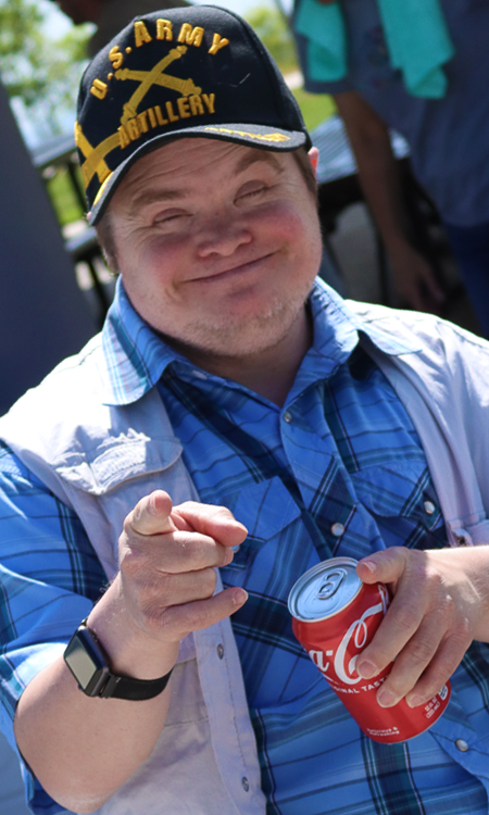 Man in blue plaid shirt and U.S. Army Artillery cap smiling and holding a Coca-Cola can