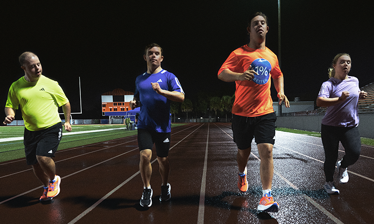 Four runners move down a track at night under stadium lights, running side by side in athletic clothing.