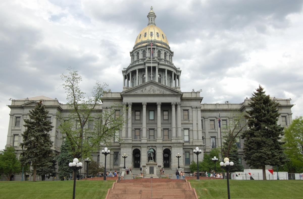 The Colorado State Capitol building stands with its gold-domed roof under a cloudy sky. The front features tall columns a grand staircase and a statue. Green trees and lampposts line the walkway.