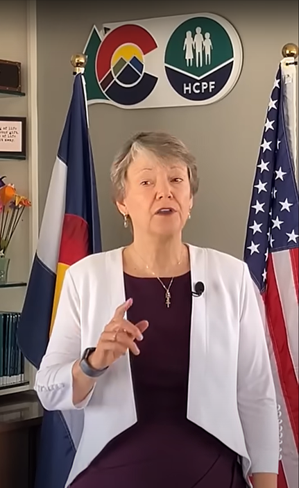 Woman stands speaking between Colorado and United States flags with HCPF logo on wall