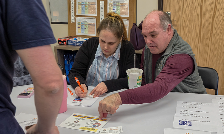 A woman writes on a worksheet while a man points to information on the table