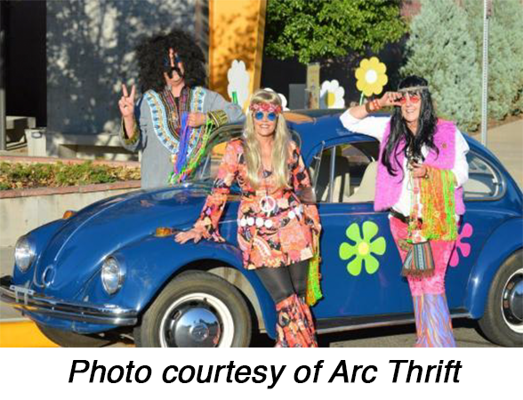 Three people in colorful 1960s-style outfits pose beside a blue Volkswagen Beetle decorated with flower decals. Text reads, â€œPhoto courtesy of Arc Thrift.â€