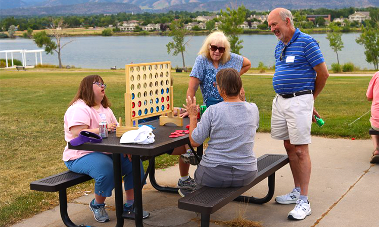 Four adults enjoy a lakeside picnic playing giant Connect Four at a table with green trees and homes in the background under a cloudy blue sky