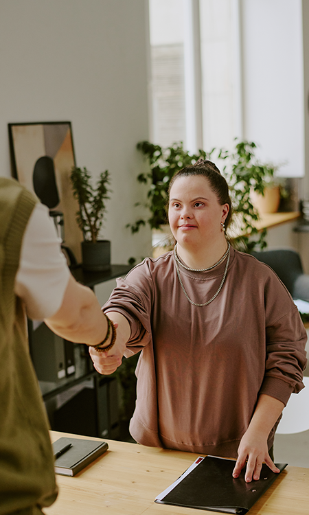 A young adult with Down syndrome shakes hands with another person across a desk in an office setting.