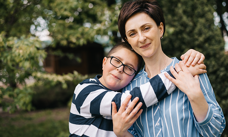 A child wearing glasses and a striped shirt hugs a woman outdoors as she holds his arm.