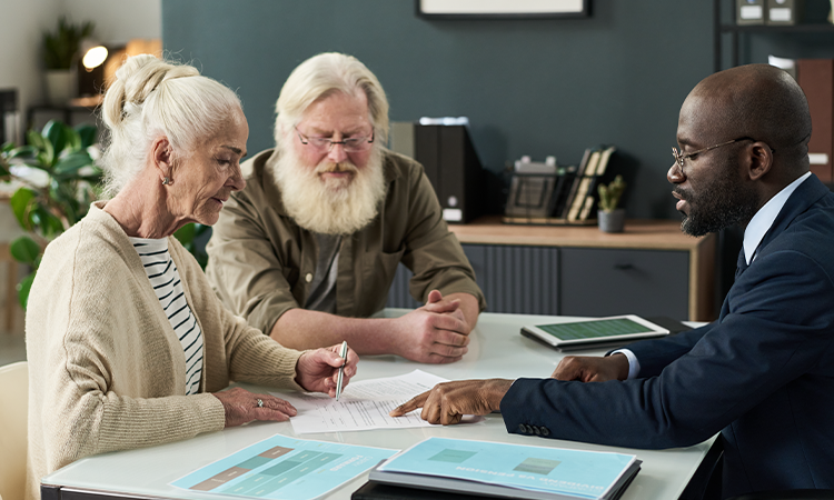 An older couple sits at a desk reviewing documents with a professional in a suit who points to paperwork while explaining details