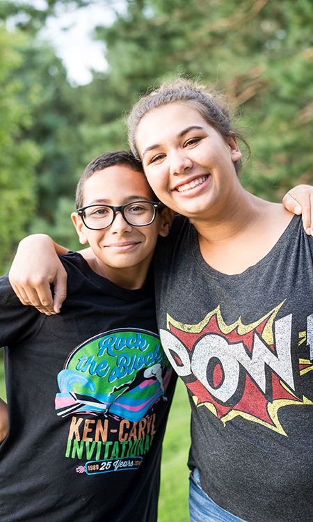 Two young people smile with their arms around each other outdoors.