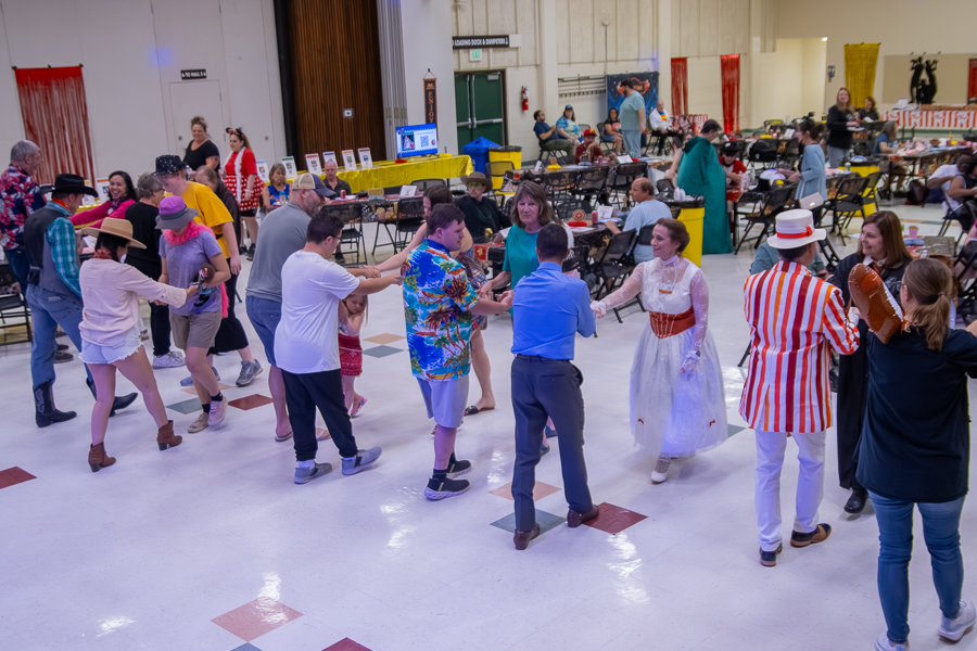People dance together in a large decorated event hall while others sit at tables in the background. Several attendees are wearing colorful or themed outfits.