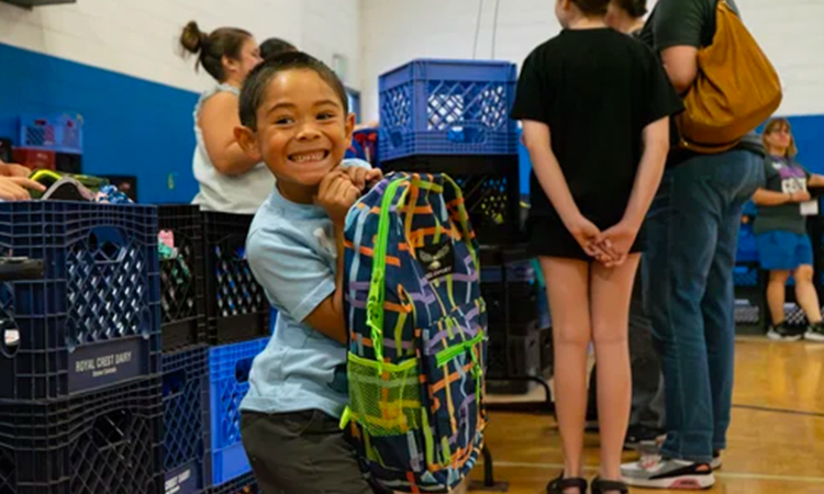 A smiling child holds a colorful backpack beside stacks of plastic crates in a school gym, with people in the background.