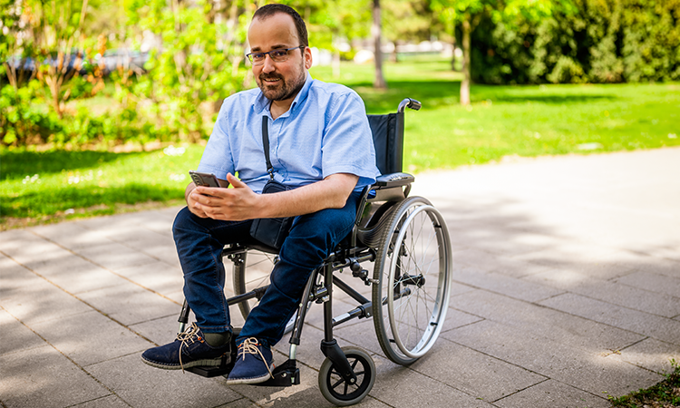 A man using a wheelchair looks at his phone while sitting on a paved path in a park.