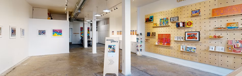 The image shows a modern art gallery with white walls and a polished concrete floor. Colorful artwork is displayed on pegboard walls and framed pieces on the left. A wooden sign in the center reads Access Gallery with an arrow pointing forward.