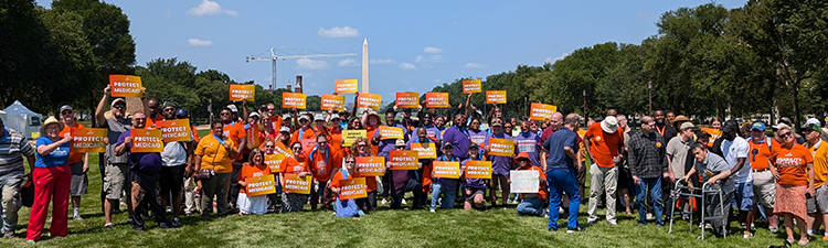 A large group of people gather outdoors holding bright orange signs that read Protect Medicaid with the Washington Monument visible in the background