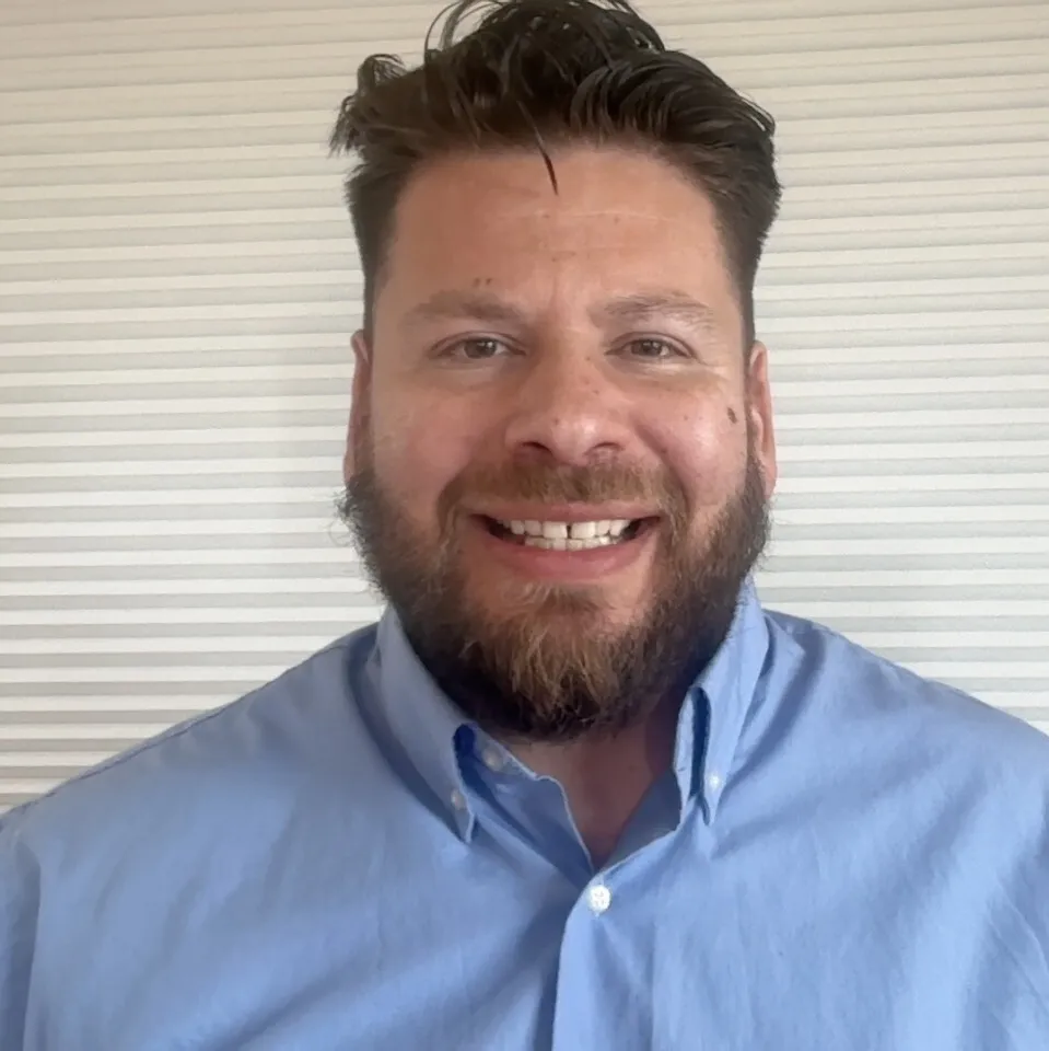 Head-and-shoulders portrait of a smiling bearded man wearing a light blue button-down shirt against a white textured background.