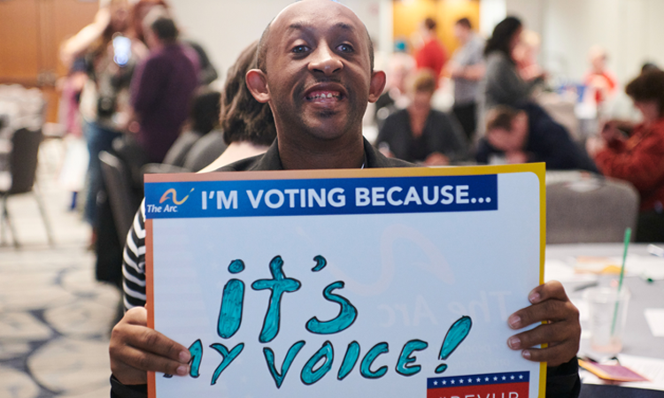 A person holds a sign reading, âIâM VOTING BECAUSEâ¦ itâs my voice!â at a community event.