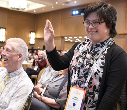 A woman wearing glasses and a conference badge raises her hand while smiling beside seated attendees at a convention.
