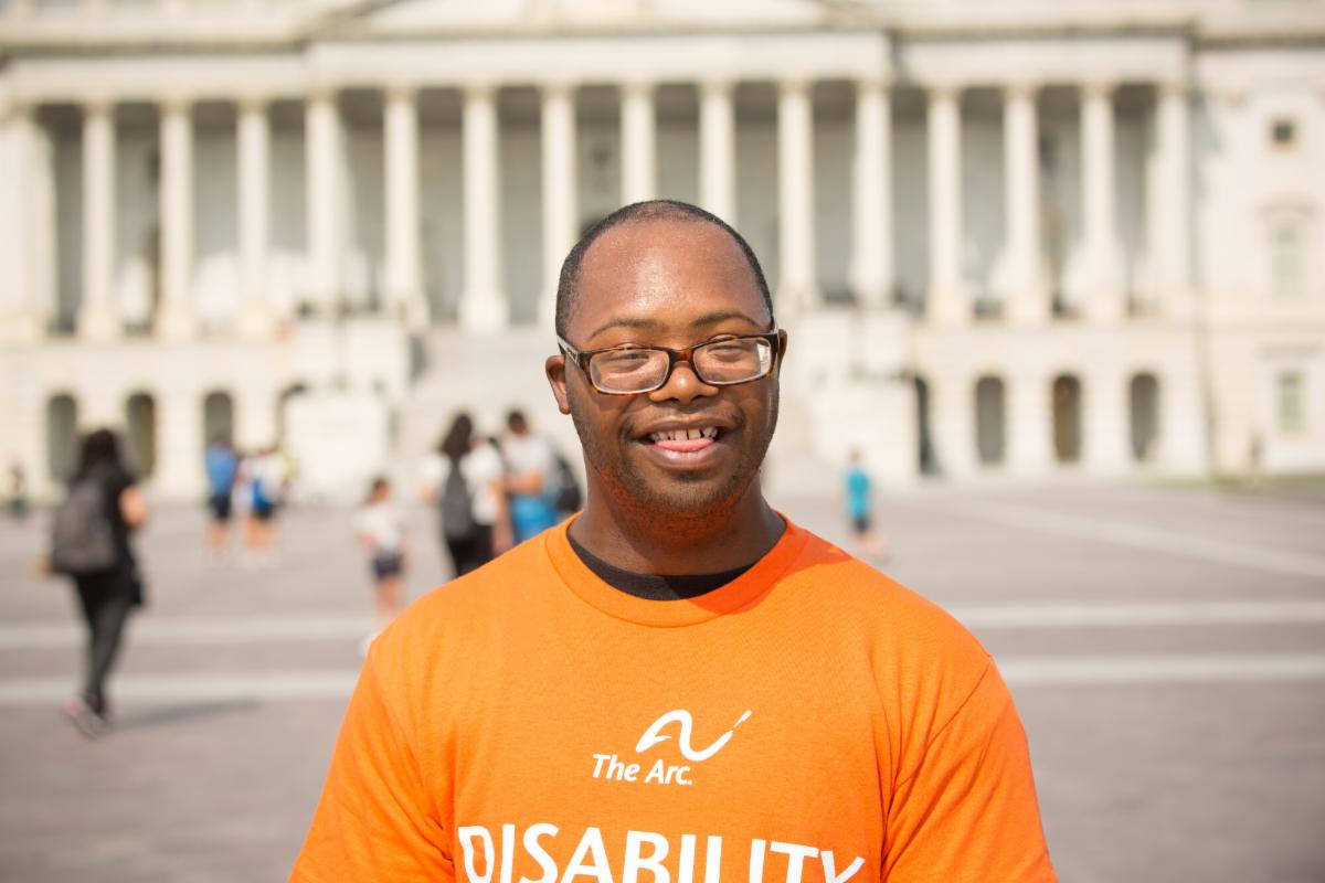 A person wearing an orange The Arc T shirt with the word disability stands smiling in front of a large white building with columns