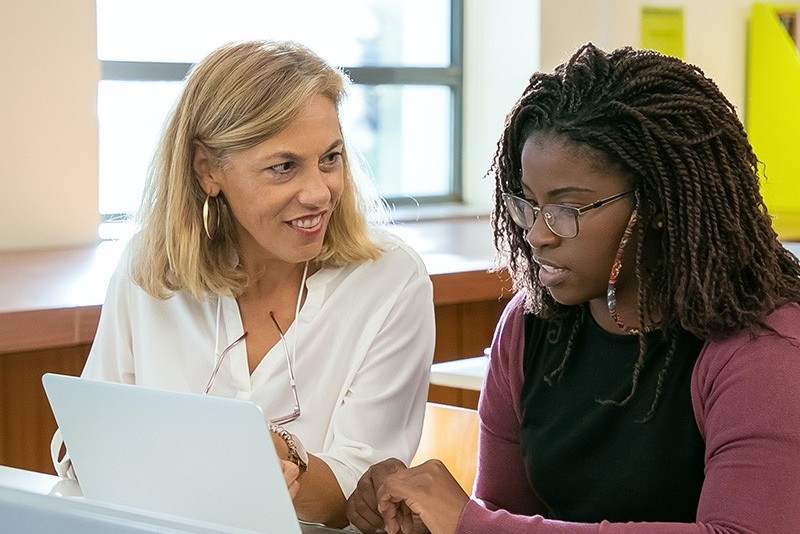Two people sit together at a table looking at a laptop, with one pointing at the screen while they talk.