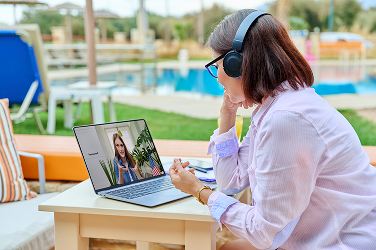 Woman talking to another woman on a computer. She sits outside beside a pool and wear headphones.