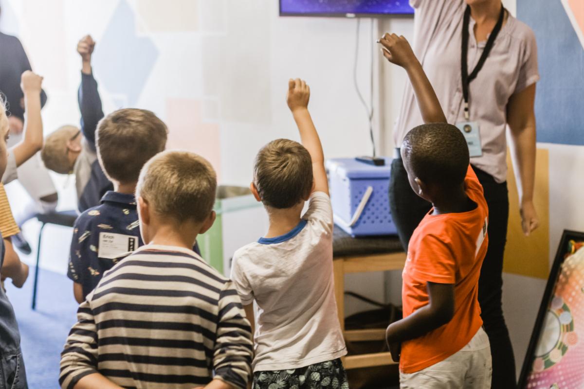 The image shows young children in a classroom raising their hands. They face an instructor who is also raising a hand. The children wear different casual clothing. The background has a mounted screen and a blue storage container on a table.