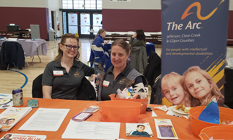 Two women smiling at a booth in a gym wearing gray shirts with logos The booth has orange baskets pamphlets and a banner reading The Arc Jefferson Clear Creek and Gilpin Counties for people with intellectual and developmental disabilities.