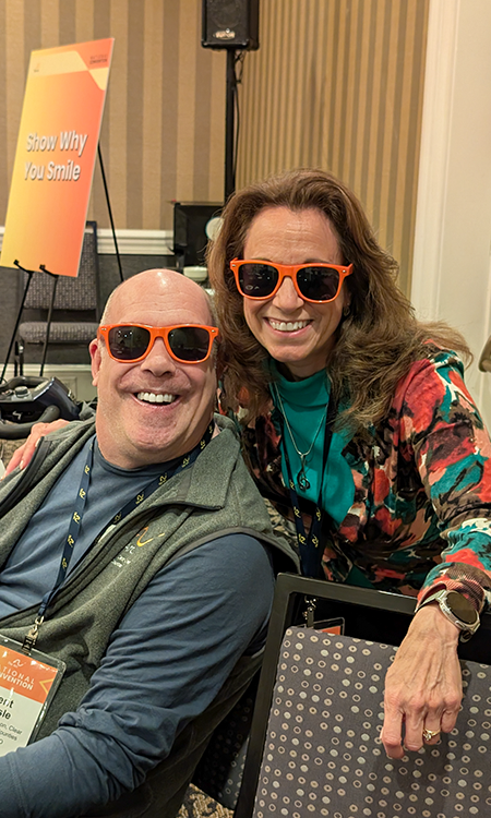 Two people smiling and posing together indoors, both wearing bright orange sunglasses. The man is seated and the woman stands beside him with her arm around his shoulder. A sign in the background reads âShow Why You Smile.â